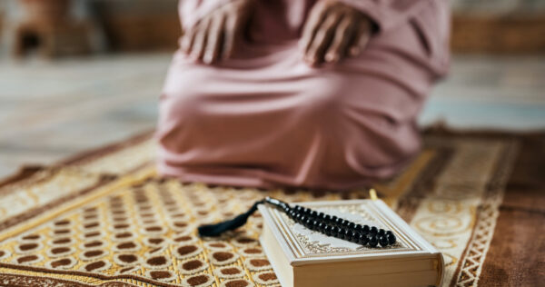 Close-up of holy book Koran and prayer bead with Muslim woman kneeling in the background.